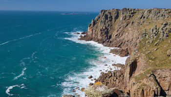 Longships lighthouse cliffs wide This landscape photograph shows the Longships lighthouse cliffs along the coast of Cornwall, United Kingdom. The image, taken in the afternoon during spring, captures the rugged cliffs meeting the clear blue waters of the Atlantic Ocean. The Longships lighthouse is visible in the distance, sitting on a small rocky islet offshore, and the natural formations and textures of the cliffs highlight the dramatic nature of this stretch of coastline in Cornwall.
